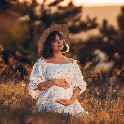 Happy Mother Carrying Child, Expecting For Baby. Amazing Motherhood. Pregnant Woman in beautiful dress and straw hat sitting on grass at nature rural meadow. High quality