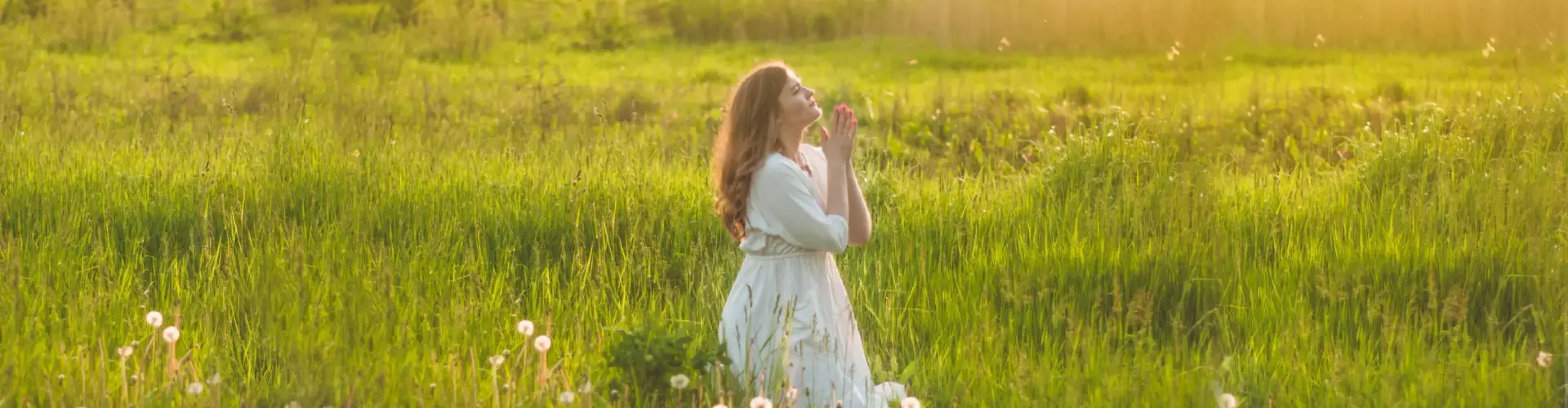 Girl closed her eyes, praying in a field during beautiful sunset. Hands folded in prayer concept for faith, spirituality and religion. Peace, hope, dreams concept